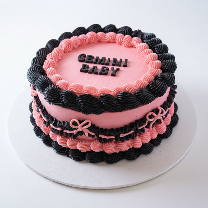 A two layer round cakette with pink and black buttercream, decorated with the words 'GEMINI BABY' on top in bold letters.