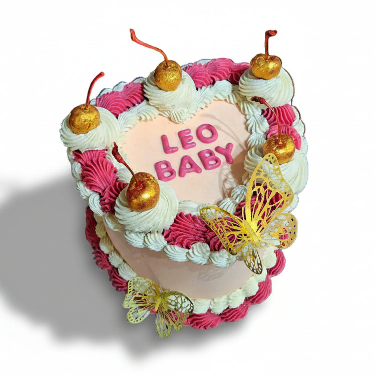 Heart-shaped cakette with 'LEO BABY' text, pink and white buttercream, gold butterflies and cherries.